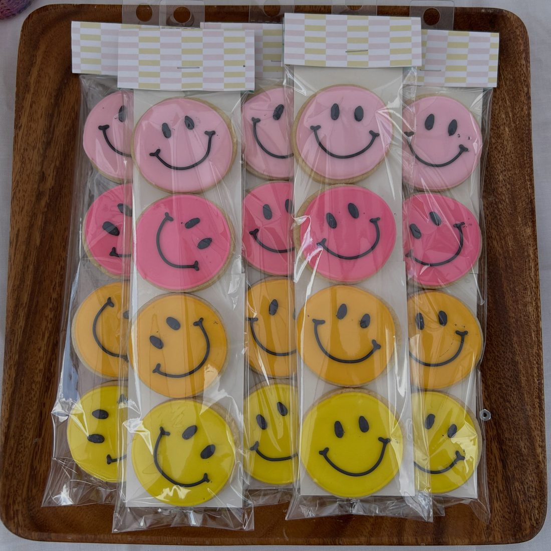 Pink, orange and yellow smiley face cookies in clear bags on a wooden tray